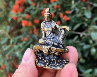 May include: A small, bronze-colored statue of a seated figure with ornate details. The figure is seated on a textured base with swirling patterns. The statue has a serene expression and is set against a blurred background of green foliage and red flowers.