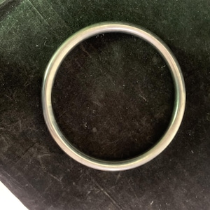 May include: A silver-coloured, circular bangle bracelet. The bracelet is made of a smooth, shiny metal and has a simple, minimalist design. The bangle is shown against a dark, textured background.