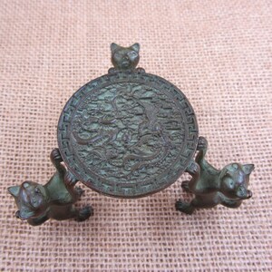 Könnte beinhalten: Ein bronzefarbener dekorativer Ständer mit einer kreisförmigen Oberseite mit einem komplizierten Drachen- und Phönix-Design. Der Ständer wird von drei Katzenfiguren getragen, eine Katze oben und zwei, die die kreisförmige Oberseite halten.
