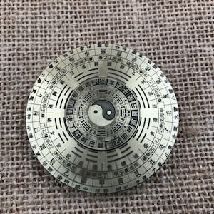 Könnte beinhalten: Ein runder, messingfarbener Kompass mit komplizierten chinesischen Schriftzeichen und einem Yin-Yang-Symbol in der Mitte. Der Kompass ist mit konzentrischen Kreisen und einem labyrinthartigen Muster gestaltet, vor einem strukturierten, beigen Hintergrund.