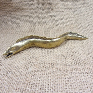 May include: A brass eel figurine with an open mouth and visible teeth. The eel is in a curved position, with a textured surface. The figurine is a golden color and appears to be made of metal.