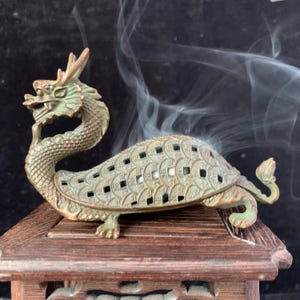 Puede incluir: Un quemador de incienso de color bronce en forma de dragón-tortuga. La dragón-tortuga se asienta sobre una base de madera, con humo que se eleva desde la parte superior. La dragón-tortuga tiene un diseño detallado con recortes en forma de moneda en su caparazón.