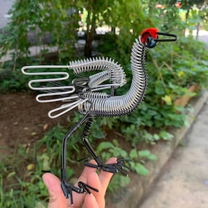 以下が含まれることがあります： 赤い頭、黒い嘴、銀色の翼を持つ鳥のワイヤー彫刻。 鳥の体はコイル状のワイヤーでできており、足は黒色です。 彫刻は、緑の葉を背景に屋外に展示されています。