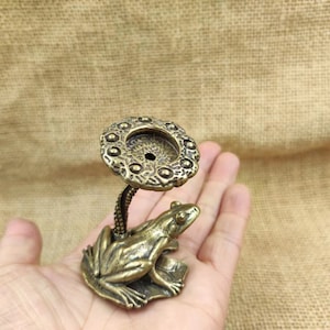 May include: A bronze frog ornament with a decorative, circular top. The frog sits on a leaf, and the top is supported by a curved stem. The frog and top have an aged, antique appearance.