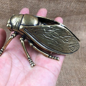 May include: A detailed bronze-colored cicada sculpture. The insect has intricate wing and body details, with six legs and large eyes. The sculpture is held in a hand, showcasing its size and craftsmanship. The cicada is a decorative item.