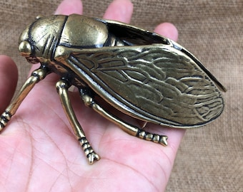 May include: A detailed bronze-colored cicada sculpture. The insect has intricate wing and body details, with six legs and large eyes. The sculpture is held in a hand, showcasing its size and craftsmanship. The cicada is a decorative item.
