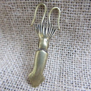 Könnte beinhalten: Ein ohrring in Form eines Tintenfischs in Messingfarbe mit einem Haken. Der Ohrring hat ein detailliertes Design eines Tintenfischs, einschließlich Tentakeln und einem Körper. Der Haken ist gebogen und oben am Tintenfisch befestigt.
