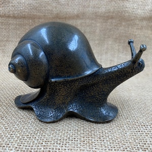 May include: A bronze-colored snail sculpture with a textured body and a smooth, rounded shell. The snail's antennae are extended, and the sculpture has a detailed, lifelike appearance. The snail is a decorative item.