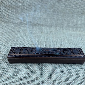 May include: A rectangular, dark brown incense burner with intricate floral and geometric patterns. Smoke rises from the burner, indicating use. The burner sits on a textured, neutral-toned surface, suggesting a natural aesthetic.
