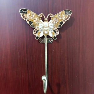 May include: A gold-tone butterfly-shaped hook with a pearl accent. The butterfly has brown and white wings with rhinestones. The hook is attached to a gold-tone metal rod.