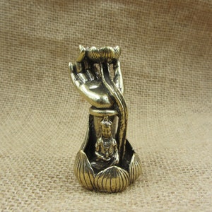 May include: A brass-colored sculpture of a hand holding a lotus flower, with a small Buddha figure seated inside the flower's base. The sculpture has intricate details and a vintage aesthetic, set against a neutral background.