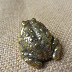 May include: A small, antique-style brass frog figurine. The frog has detailed, textured skin and a slightly open mouth. The brass has a warm, aged patina, giving it a vintage appearance. The frog is a decorative item.