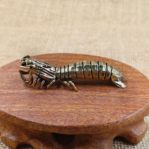以下が含まれることがあります： 詳細なカマキリの真鍮製フィギュア。彫刻は金色がかった茶色で、小さくて丸い木製の台の上に置かれています。カマキリは、体と爪が細かく分かれており、非常に詳細です。