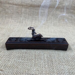 May include: A bronze-colored incense burner with a rectangular base and a decorative deer figurine on top. The base has intricate patterns, and the deer is positioned above several coins. Smoke gently rises from the burner, suggesting its use.