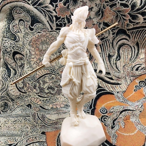 Może przedstawiać: Kremowa figurka wojownika ze złotą laską. Postać stoi na małej podstawie, z dopracowanymi detalami odzieży i definicją mięśni. Tło ma wzorzysty deseń.