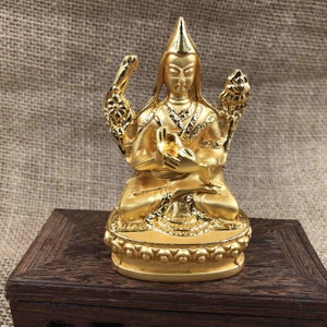 May include: A gold-colored statue of a seated figure with a pointed hat and ornate detailing. The figure is in a meditative pose, with hands held in a gesture of prayer. The statue is on a dark wooden base, set against a neutral background.
