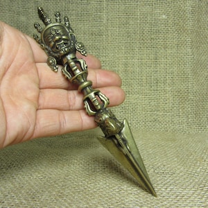 May include: A bronze-colored ritual dagger with intricate details. The handle features a detailed face and hand design, while the blade is a sharp, triangular shape. The dagger is held in a hand against a neutral background.