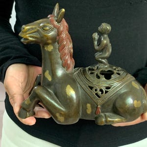 Puede incluir: Quemador de incienso de bronce con forma de caballo reclinado con un mono en la espalda. El caballo tiene una melena roja y detalles dorados. El quemador de incienso tiene un diseño calado y ornamentado y lo sostiene una persona.