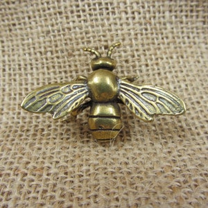 May include: A detailed, antique-style brass bee brooch with outstretched wings. The bee has intricate wing detailing and a textured body, showcasing a vintage aesthetic. The brooch is a decorative accessory.