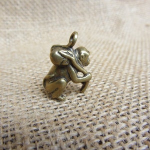 May include: A small, bronze-colored monkey figurine. The monkey is depicted in a squatting position, carrying a sack on its back and holding a curved object. The figurine has a loop at the top for hanging. The overall design is detailed and textured.