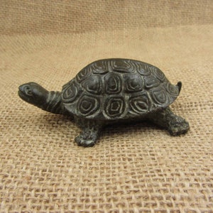 May include: A bronze-colored turtle figurine with detailed shell patterns and visible legs and head. The sculpture is a decorative item, likely for home decor or a collector's piece. The turtle is positioned on a neutral-toned surface.