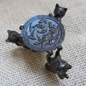 Könnte beinhalten: Ein bronzefarbener dekorativer Ständer mit drei Katzenfiguren, die eine kreisförmige Platte tragen. Die Platte hat ein geprägtes Blumendesign und einen dekorativen Rand. Die Katzen sind um den Sockel herum positioniert und halten die Platte hoch.
