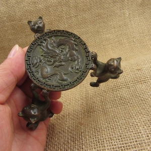 Könnte beinhalten: Bronze-farbenes dekoratives Objekt mit einem kreisförmigen Design mit einem Drachenmotiv. Drei kleine katzenartige Figuren sind um den Kreis herum positioniert und scheinen ihn zu halten. Das Objekt hat ein gealtertes, antikes Aussehen.