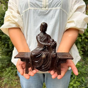 May include: A dark brown sculpture of a seated figure with a smooth, polished finish, wearing traditional robes with intricate etched patterns. The figure rests on a rectangular bench with carved legs and a wood-grain texture on the surface.