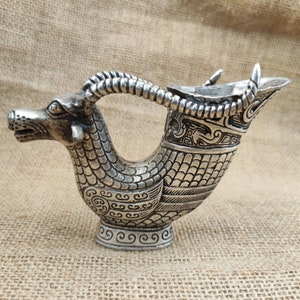 May include: A silver-colored decorative vessel with a ram's head and curved handle. The vessel features intricate engraved patterns and a spout. The base has a decorative swirl design. The background is a tan burlap.