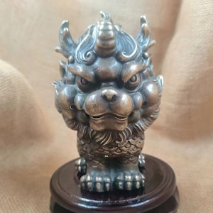 Könnte beinhalten: Eine detaillierte, bronzefarbene Skulptur einer mythischen Kreatur mit löwenartigem Gesicht, Hörnern und einer strukturierten Mähne. Die Skulptur ist auf einem dunklen Holzsockel montiert und zeigt aufwendige Handwerkskunst und eine Vintage-Ästhetik.