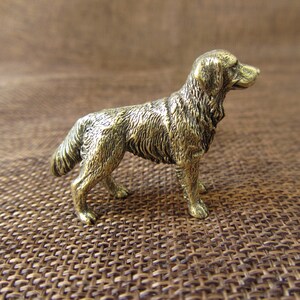 Könnte beinhalten: Kleine, detaillierte Messingfigur eines Golden Retriever-Hundes in stehender Pose. Der Hund hat ein strukturiertes Fell und blickt nach rechts. Der Hintergrund ist braun gewebt.