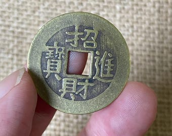 May include: A round, brass-colored metal coin featuring a square hole in the center. The surface is embossed with four Chinese characters arranged around the square opening, specifically the characters 招, 進, 寶, and 財.