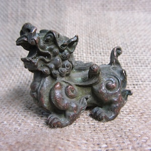 May include: A small, bronze-colored statue of a mythical creature, possibly a Foo Dog or guardian lion. The figurine has detailed features, including a snarling expression, curled mane, and ornate detailing. The statue is approximately 3 inches long.