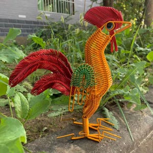 Op de afbeelding: Een handgemaakte metalen haan sculptuur. De haan heeft een rode kam en staart, een oranje lichaam en groene accenten. De sculptuur is gemaakt van draad en staat op een betonnen oppervlak, omringd door groen. De haan is een decoratief tuinornament.