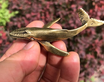 May include: A small, brass whale figurine with detailed fins and tail. The whale is a golden color and appears to be made of metal. The figurine is held in a hand against a blurred background of red and green foliage.