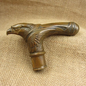 Puede incluir: Empuñadura de bastón de color bronce con diseño de cabeza de águila. La cabeza del águila está detallada, con plumas y un pico afilado. El mango es curvo y tiene una zona de agarre texturizada. Empuñadura de bastón de metal.