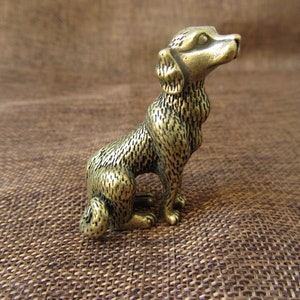 Puede incluir: Pequeña figura de perro de latón de estilo antiguo, sentado con la cabeza inclinada hacia arriba. El perro tiene una textura de pelaje detallada y una apariencia ligeramente envejecida, lo que sugiere que es un artículo vintage o artesanal. Es una pieza decorativa.