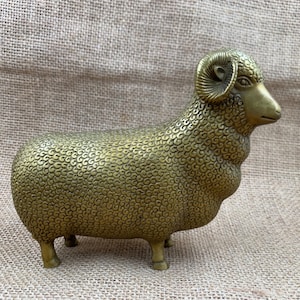 May include: A brass-colored ram sculpture with detailed textured wool and curved horns. The ram stands on four small legs, with a detailed face and a slightly open mouth. The sculpture is a decorative item.