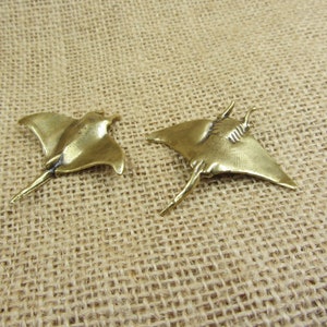 Può includere: Due statuette di manta color bronzo. Le razze hanno pinne a forma di ali e lunghe code. Le statuette sono esposte su una superficie strutturata e beige, che suggerisce un ambiente naturale o rustico. Le razze sono rivolte in direzioni diverse.