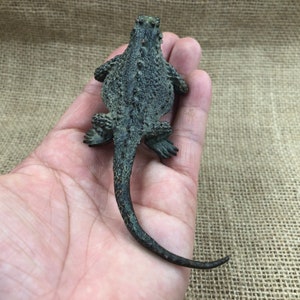 May include: A detailed, bronze-colored lizard figurine with textured skin and a long tail. The figurine is lying on its back, showcasing its intricate design and realistic features. The lizard's legs are positioned as if it is crawling.