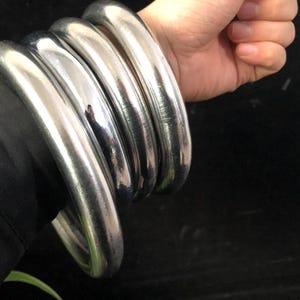 May include: Four silver-coloured, thick metal bangles. The bangles are smooth and shiny, reflecting light. The jewellery is worn on a wrist, with the hand visible.