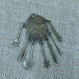 Könnte beinhalten: Ein detaillierter Anhänger im antiken Stil mit einem zentralen kreisförmigen Design mit einem komplizierten Muster und einem Yin-Yang-Symbol. Der Anhänger ist mit verschiedenen Miniaturwaffen-Anhängern verziert, darunter Schwerter, ein Dreizack und eine Axt. Das Metall hat ein angelaufenes, gealtertes Aussehen.