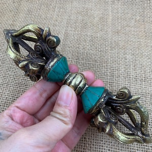 May include: A brass vajra ritual object featuring intricate scrollwork and two inlaid turquoise-coloured segments. The central handle is ribbed, flanked by two conical sections with teal stone inlays, and ends in ornate prongs.