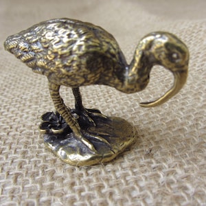 Peut inclure: Petite figurine d'oiseau de couleur bronze, avec un bec courbé et un long cou fin. L'oiseau est posé sur une base en forme de feuille avec une petite fleur. La figurine a une surface texturée et détaillée, probablement en métal.