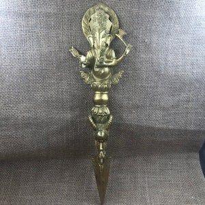 Könnte beinhalten: Ein verzierter Dolch in Messingoptik, der die hinduistische Gottheit Ganesha darstellt. Der Dolch hat eine spitze Klinge und einen Griff mit aufwendigen Schnitzereien, darunter elefantenköpfige Figuren und dekorative Elemente.