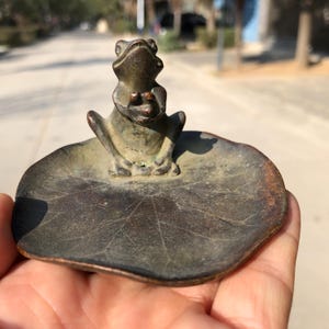 May include: A bronze frog figurine sitting on a lily pad-shaped dish. The frog is in a seated position with its hands clasped. The dish has a textured surface and a dark, aged appearance. The overall design is detailed and artistic.