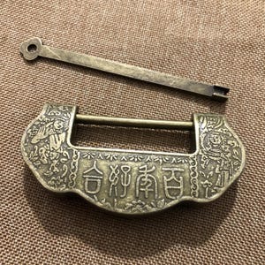 Puede incluir: Un candado antiguo de color bronce con una llave a juego. El candado tiene intrincadas tallas y caracteres chinos. La llave es una barra larga y delgada con un agujero circular en un extremo. El candado es rectangular con la parte superior e inferior curvadas.