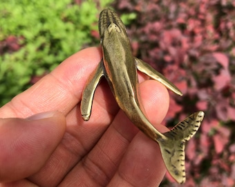 May include: A detailed brass whale figurine with fins and a tail. The whale is a golden color and has a textured surface. The figurine is held in a hand against a background of green and red foliage.