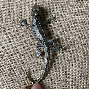May include: A metallic bronze-toned lizard figurine featuring a textured, scaly surface across its back and head. The lizard is depicted in a crawling position with four legs extended outward and a long, curved tail extending towards the bottom edge.