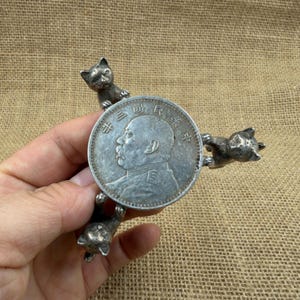 Puede incluir: Una moneda de plata con un retrato y caracteres chinos, sostenida por tres pequeñas y detalladas figuras de gato. La moneda es redonda y tiene un diseño detallado. Los gatos están colocados alrededor de la moneda, dándole un aspecto artístico único.